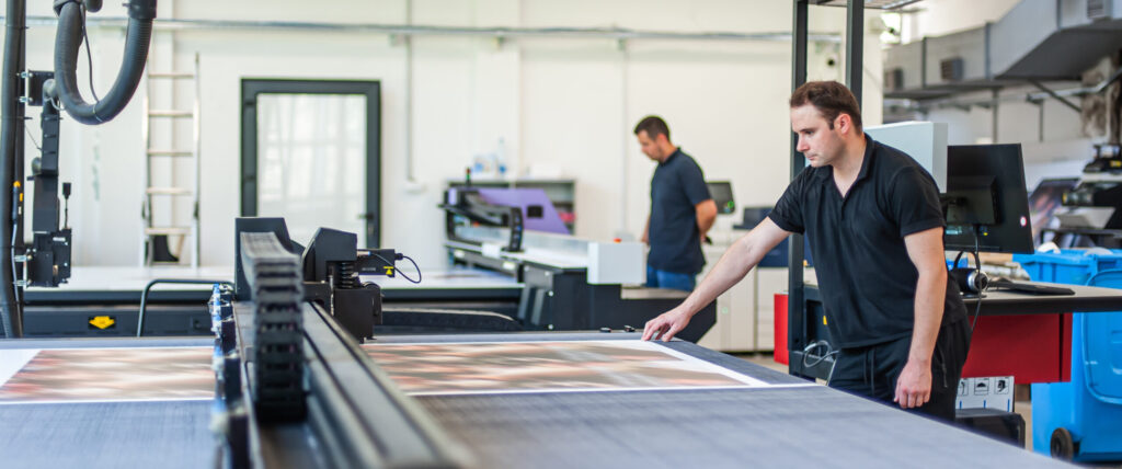 Technicians diligently manage large-format industrial machinery on a busy production floor, illustrating the critical operational errors and ways print shops lose jobs to more efficient competitors.